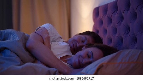 Side view of husband hugging wife falling asleep together in comfortable bed. Young happy couple lying in bed and sleeping late at night in dark bedroom at home