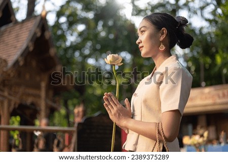 Side view image of a beautiful Asian woman in a traditional Thai-Lanna dress with a lotus flower in her hands is making a wish in a temple. praying, paying respect to the Buddha, Thai culture