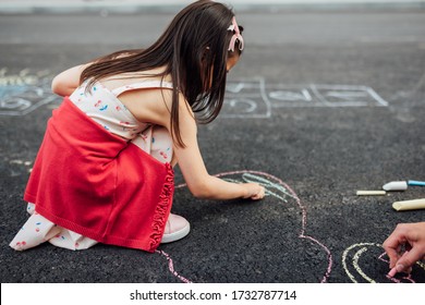 Side view image of little girl drawing with colorful chalks on playground. Child playing the summer's games outside. Kid drawing with chalk a heart on grey pavement. Activities for children.
