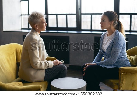 Side view image of middle aged female psychologist discusses the problems with young pregnant woman, sitting in armchairs, facing each other. Prenatal psychological therapy