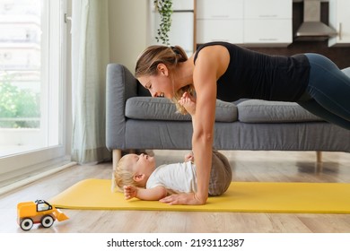 Side View Image Of Mother Practicing Yoga Standing In Plank Position Above His Cute Baby Boy Lying On Mat, Reaching His Legs To Her Face. Mom Working Out With Her Toddler. Happy Healthy Motherhood