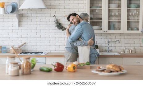 Side view image of a young adult caucasian son hugging embracing showing his care and love to elderly old senior father while cooking meal, food in the kitchen. Happy father`s day! I love you, dad! - Powered by Shutterstock