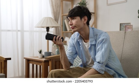 Side View Of Impatient Asian Man Shaking Head With Dissatisfied Look On Face While Channel Surfing With A Tv Controller On The Couch In The Living Room At Home