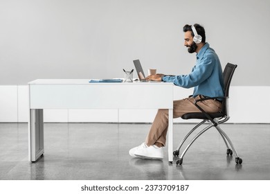 Side view of indian businessman in headphones working on laptop, making video call, sitting at desk, having web conference with customer in office, free space