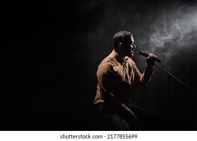 Side View Of Indian Comedian Sitting On Chair And Performing Stand Up Comedy Into Microphone On Black With Smoke