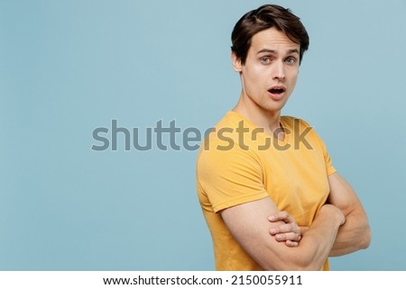 Side view indignant young man 20s wear yellow blank print design t-shirt hold hands crossed folded look camera isolated on plain pastel light blue background studio portrait. People lifestyle concept