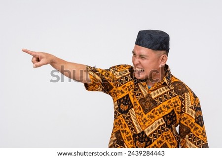 Side view of an indonesian man in traditional batik shirt and kopiah hat visibly angry, pointing and blaming someone, or making grave threats. Against a white backdrop.