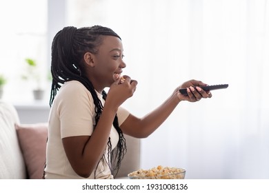 Side View Of Joyful African American Woman Watching Television And Eating Popcorn At Home, Closeup, Copy Space. Positive Black Lady Sitting On Couch In Living Room, Holding TV Remote