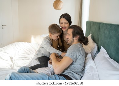 Side View Of The Joyful Happy Family Sitting Together At The Bed While Playing At Game At The Bedroom. Son Tickling His Father Laughing Out Loud. Family Great Weekend Concept