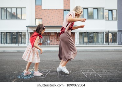 Side View Of A Joyful Little Girl Playing Hopscotch With Her Mother On Playground Outdoors. A Child Plays With Her Mom Outside. A Kid And Mum Play Hopscotch Drawn On The Pavement Outside. 