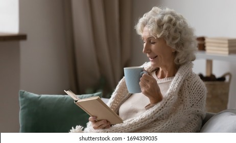 Side view joyful mature older woman covered in knitted plaid reading paper book with poetry, drinking tea. Excited middle aged elderly granny entertaining, enjoying weekend hobby time alone at home. - Powered by Shutterstock