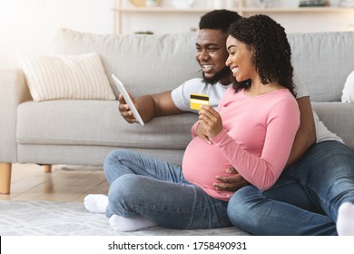 Side view of joyful pregnant black woman shopping online with husband, man holding digital tablet, sitting on floor at home, copy space