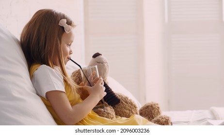 Side View Of Kid Drinking Juice Near Teddy Bear On Bed