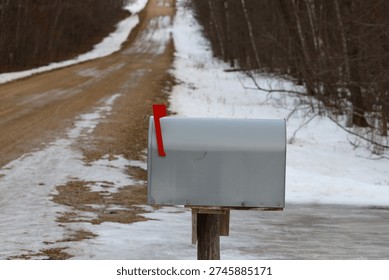 Side view of a large light gray rural mailbox with the red flag raised. A snowy winter scene shows a gravel road and bare deciduous forest trees in the background.
