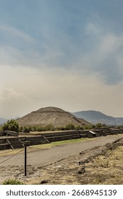 A side view of a large, stepped pyramid, showing its rounded top and tiered base, with mountains in the background and a paved walkway in the foreground