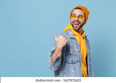 Side view of laughing young hipster guy in fashion jeans denim clothes posing isolated on pastel blue background studio portrait. People lifestyle concept. Mock up copy space. Pointing thumb aside