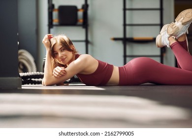 Side View. Laying Down On The Ground And Smiling. Young Beautiful Woman In Sportive Clothes Have Fitness Day In The Gym.