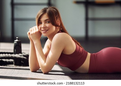 Side View. Laying Down On The Ground And Smiling. Young Beautiful Woman In Sportive Clothes Have Fitness Day In The Gym.