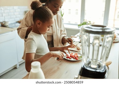 Side view of little African American girl learning how to cut strawberries for serving or making fruit smoothie and mother teaching daughter making dessert at kitchen counter, copy space - Powered by Shutterstock