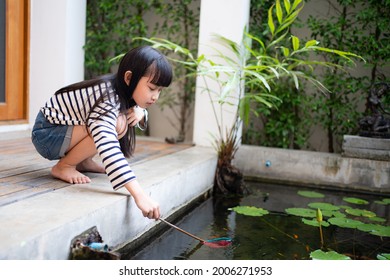 Side view of little Asian girl with long-haired and bangs squatting on floor beside the pond using a scoop net catching a small fish in the water pond with lotus leaves and plants in the home backyard