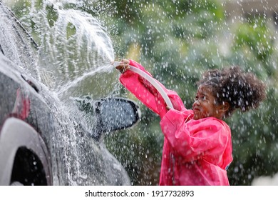 Side view of Little curly hair girl wearing raincoat colour pink playing spray water to cleaning car.