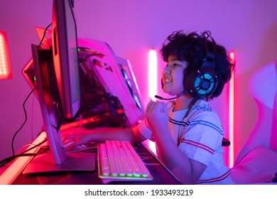 Side View Of Little Curly-haired Asian Boy In A Headset Playing Online Video Games On A Personal Computer With Right Hand Pressing On The Mouse And Happy Raising Left Fist Up After Winning The Game.