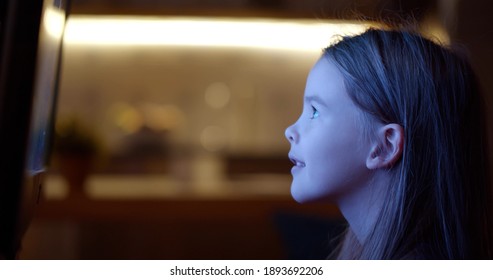 Side View Of Little Girl Sitting Neat Tv Screen And Watching Cartoons. Adorable Preteen Kid Watching Tv In Evening At Home. Kid Enjoying Movie On Television At Home