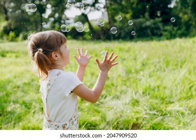 Side view little smiling happy kid girl 5-6 years old in white casual clothes blowing bubbles play on park green sunshine lawn, spending time outdoor in village countyside during summer time vacations - Shutterstock ID 2047909520