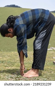 Side View Of A Long Haired Indian Young Guy Doing Uttanasana Yoga Pose In The Mountain. A Male Hiker Doing Stretching By Standing Forward Bend Toe Touch Exercise.