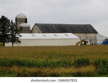 a side view of a long white outer building in front of an old grey barn and multiple silos, with tractors and trucks parked on the side and a field in the foreground
