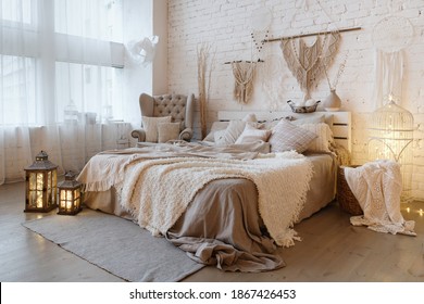Side view of lovely bedroom with plaid and pillows on comfortable bed, home decor and cushions on soft armchair in white interior design in bohemian style