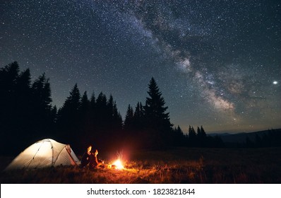 Side View Of Loving Couple Sitting Near Bright Burning Campfire And Tent, Enjoying Beautiful Camping Night Together Under Dark Sky Full Of Shiny Stars And Bright Milky Way, Warm Summer Night.