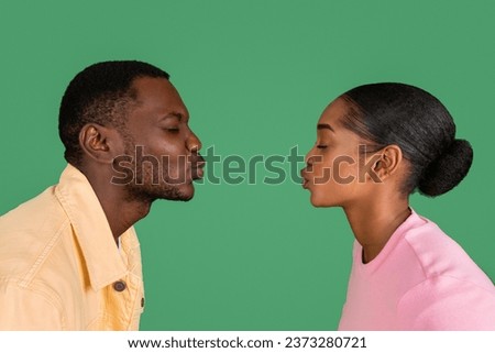 Side view of loving young black man and woman kissing with closed eyes, copy space between them, isolated on green studio background. Love, affection, bonding, relationships