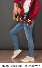 Side view of lower half of an unrecognizable female guitarist playing six string acoustic guitar. Stylish youthful musician is wearing in red leather jacket, blue jeans and sneakers