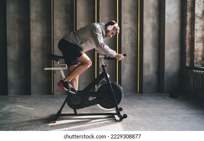 Side view of male athlete in sportswear and wireless headphones working out with stationary exercise cycle in spacious gym in daytime