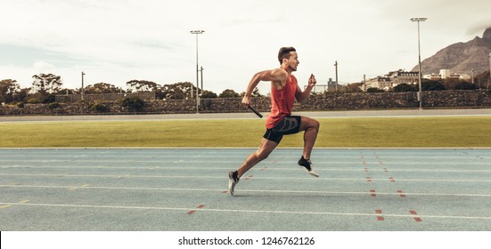 Side View Of A Male Athlete Sprinting On A Running Track In A Track And Field Stadium Holding A Baton. Male Runner Training On A Running Track.