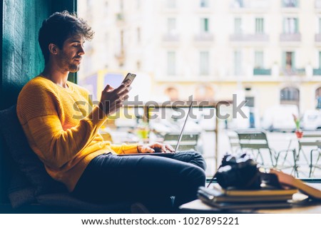 Side view of male blogger browsing profile in networks on smartphone via 4G internet connection resting in stylish coffee shop with modern laptop computer.Young man chatting online on mobile phone