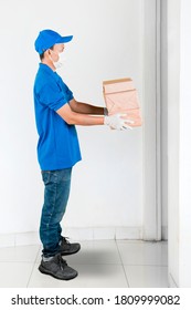 Side view of male courier wearing face mask and gloves while giving a stack of packages to customer and standing near an opened door
