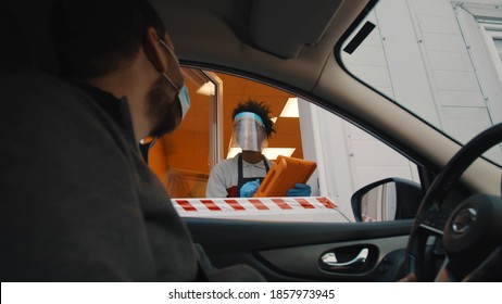 Side view of male driver in safety mask ordering meal in drive thru window. Fast food worker in face shield with tablet taking order from driver in delivery window