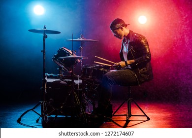 Side View Of Male Musician In Leather Jacket Playing Drums During Rock Concert On Stage With Smoke And Spotlights