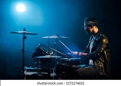 Side View Male Musician In Leather Jacket Playing Drums During Rock Concert On Stage With Smoke And Spotlight