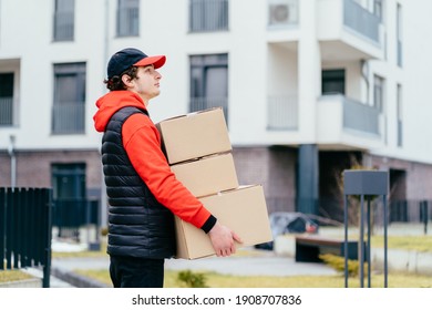 Side view of male in red uniform holding paper boxes for parcels, close-up. Light grey background with apartment house, place to insert your text. Delivery man. - Powered by Shutterstock