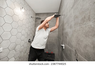 Side view of male worker using electric drilling machine while working on bathroom renovation. Man drilling wall with cordless power drill while installing shower system at home.