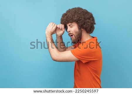 Side view of man with Afro hairstyle wearing orange T-shirt making glasses shape, looking through monocular gesture with amazed expression. Indoor studio shot isolated on blue background.