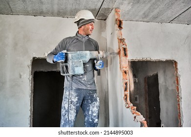 Side view of man builder in workwear drilling wall with hammer drill. Male worker using drill breaker while destroying wall in apartment under renovation. Demolition work and home renovation concept.
