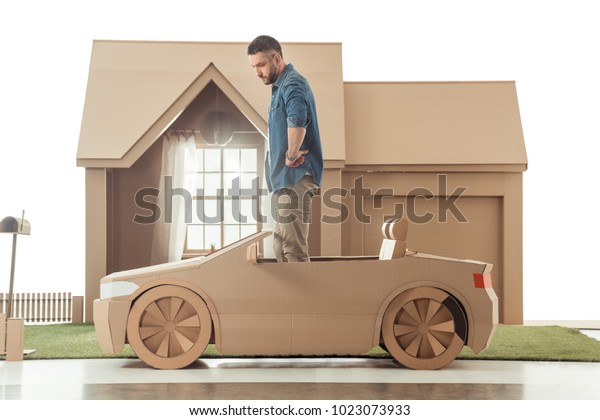 side view of man in cardboard car in front of
cardboard house isolated on
white