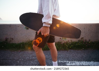 Side view of man carry longboard in his hand. Casual dressed young man in grey hoodie walk with skateboard in sunset light with leaks