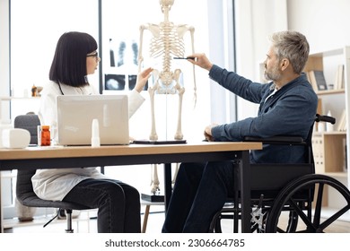Side view of man with disability asking traumatologist questions while pointing with pen at anatomical model. Female surgeon consulting patient about spinal cord injury in doctor's office at clinic.