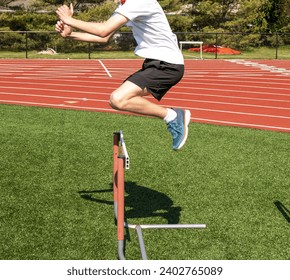 Side view of A man jumping over a hurdle on a tyrf field by a track.