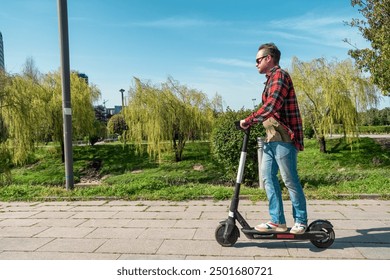 Side view of man riding e-scooter on pavement surface in city public park at sunny summer day. Male driving electric scooter outdoors. Eco-friendly city transport and travel. Sustainable lifestyle